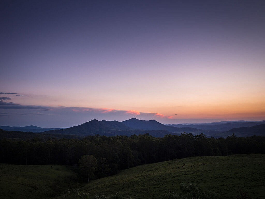 A twilight view from the edge of Border Ranges National Park, looking over dark forested ridges and rolling green farmland under a purple sky with a thin band of pink-lit clouds on the horizon.