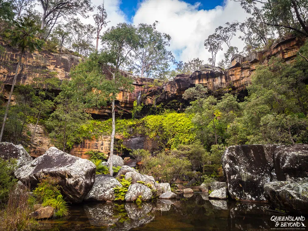 A calm, reflective rock pool at the base of Rainbow Falls (Gudda Gumoo) in Blackdown Tableland National Park, surrounded by massive granite boulders, lush ferns, and a towering sandstone cliff ledge with green vegetation.
