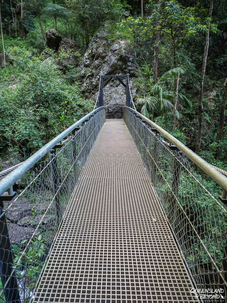 The metal-grated suspension bridge crossing the creek at the base of Purling Brook Falls. The bridge is anchored into the rocky creek bed, surrounded by lush vegetation and large basalt boulders, providing a stable crossing point for hikers.