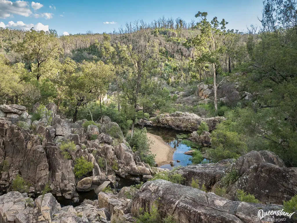 A rugged view over The Cascades, a narrow, dark creek winding through a steep granite gorge in Crows Nest National Park, surrounded by dry eucalypt forest and large grey rock formations under a clear afternoon sky.