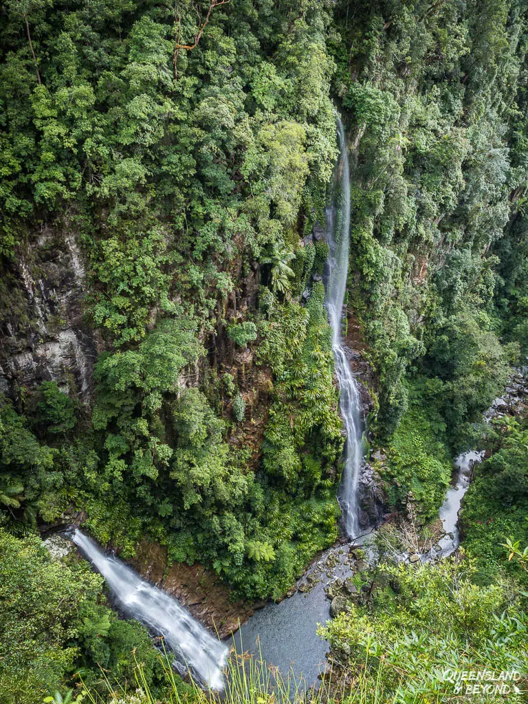 A high-angle view looking down at Coomera Falls as it plunges down a sheer, vegetation-covered cliff face into a deep rock pool below, with a second smaller waterfall joining from the left.