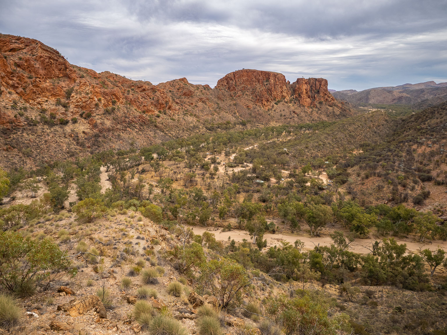 Elevated view of Trephina Gorge Nature Park in the East MacDonnell Ranges, showing a sandy, tree-lined creek bed winding through a valley flanked by rugged red rock cliffs under a cloudy sky, taken from the Panorama Walk.