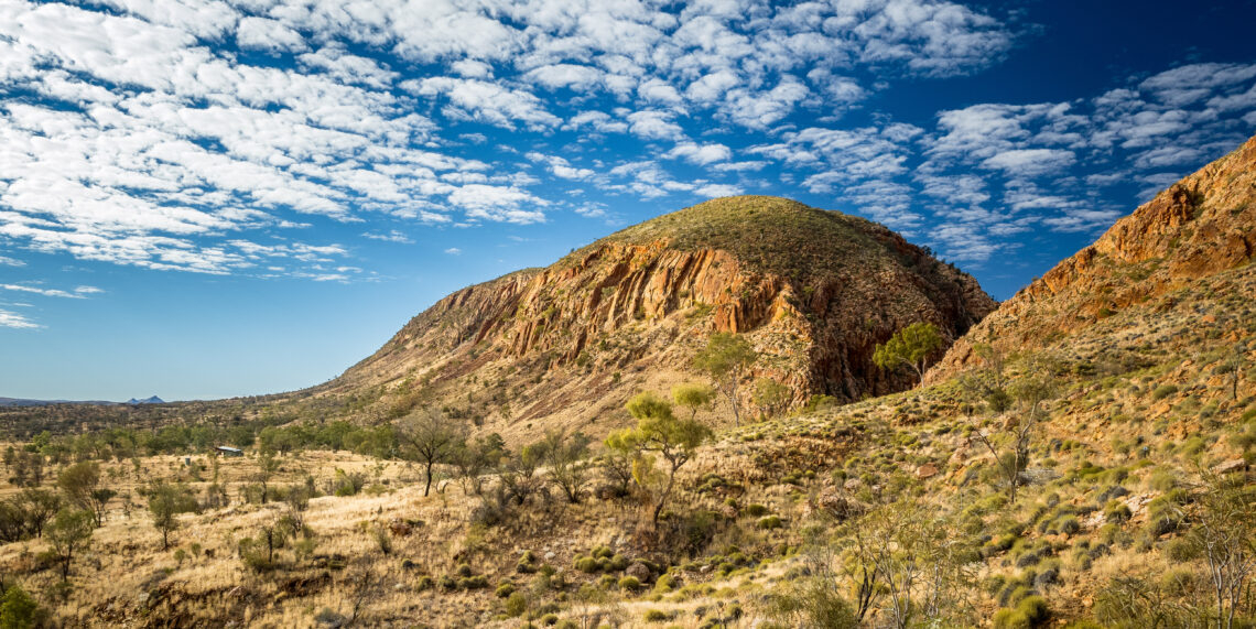 Rugged red rock cliff face of the West MacDonnell Ranges under a blue sky with textured clouds, showcasing the outback scenery of the Larapinta Trail in Australia.