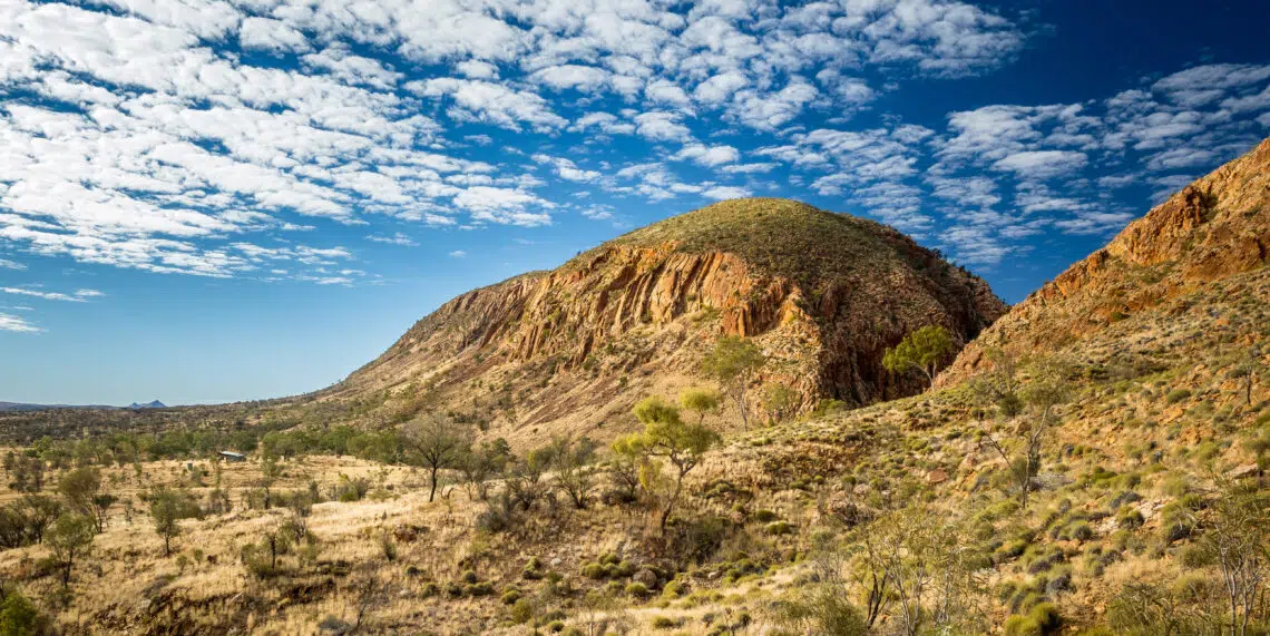 Rugged red rock cliff face of the West MacDonnell Ranges under a blue sky with textured clouds, showcasing the outback scenery of the Larapinta Trail in Australia.
