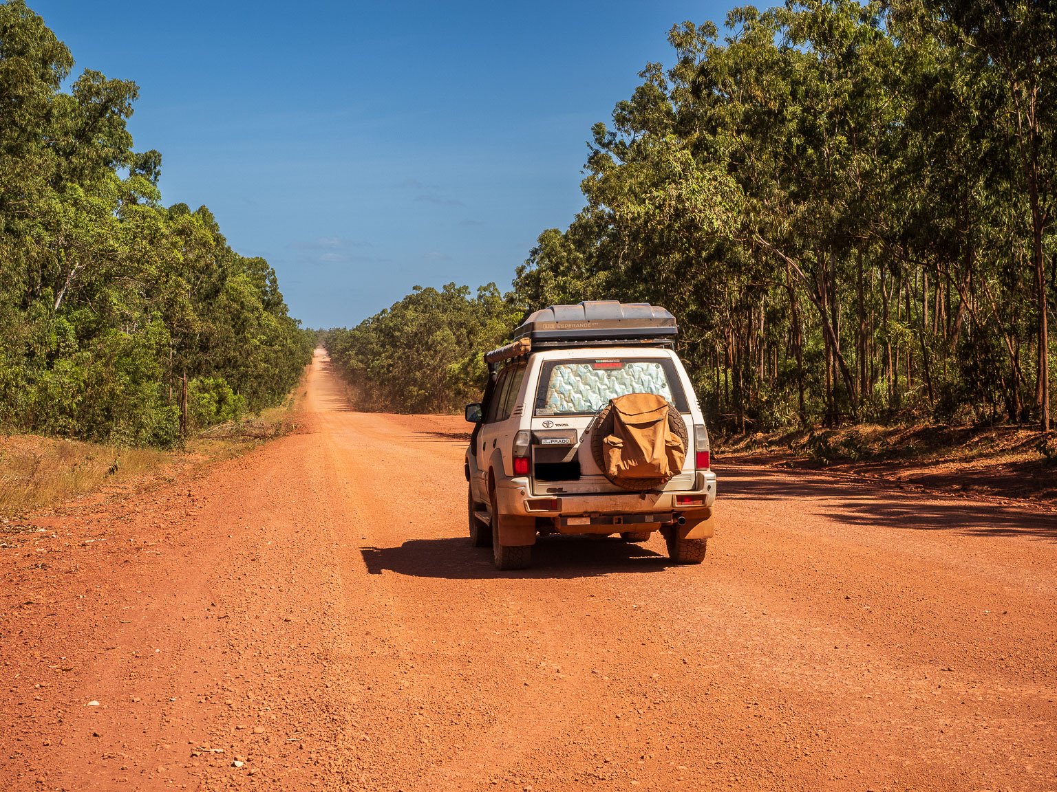 A 4WD with a rooftop tent driving down the long, red dirt Central Arnhem Road through native bushland in East Arnhem Land, Northern Territory.