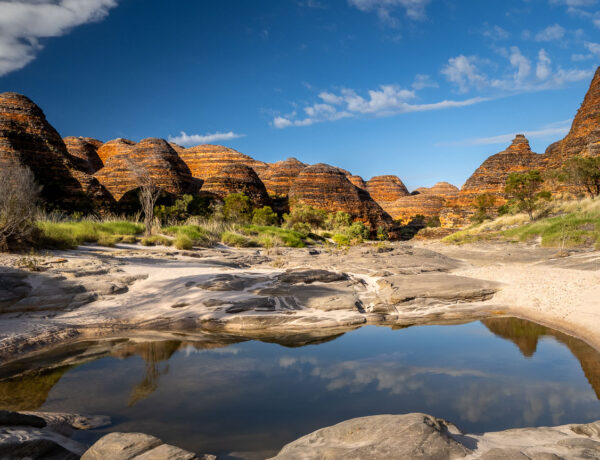A calm water reflection pool in the sandy bed of Piccaninny Gorge walking track, surrounded by the orange and black striped beehive domes of the Bungle Bungle Range in Purnululu National Park.
