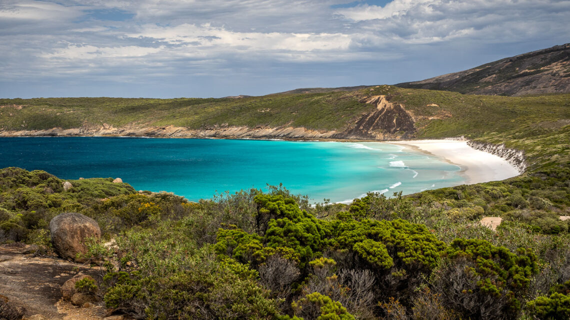 Elevated view from the Coastal Trail of the turquoise waters and white sand crescent beach of Hellfire Bay in Cape Le Grand National Park, Western Australia, framed by green coastal heath and granite rocks in the foreground under a cloudy sky.