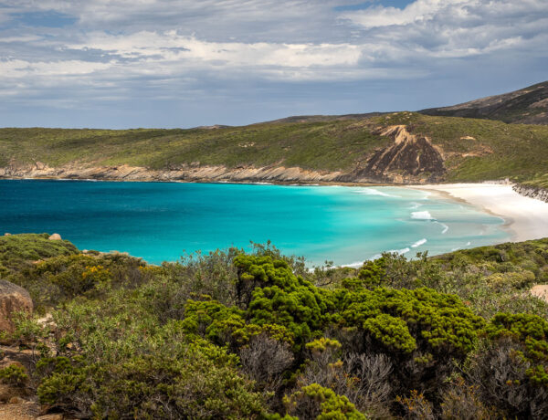Elevated view from the Coastal Trail of the turquoise waters and white sand crescent beach of Hellfire Bay in Cape Le Grand National Park, Western Australia, framed by green coastal heath and granite rocks in the foreground under a cloudy sky.