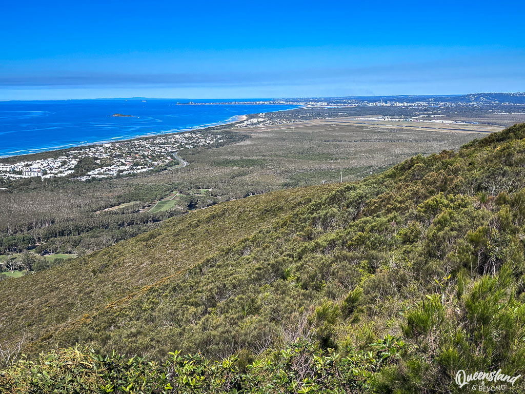 Panoramic view from the Mount Coolum walk summit on a clear winter day, looking south over a green vegetated slope towards the Mooloolaba coastline and Sunshine Coast Airport.