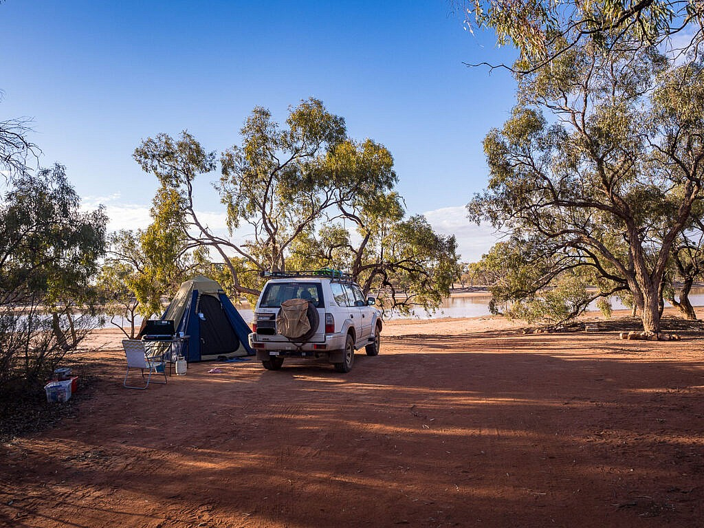 A white 4WD and a blue tent set up at a remote, sandy campsite along a lagoon. A camp chair and a fuel stove on a small table are positioned in front of the tent under the shade of leaning eucalyptus trees. The ground is rich, red Australian soil, and the calm water of the river reflects a clear blue sky in the background.